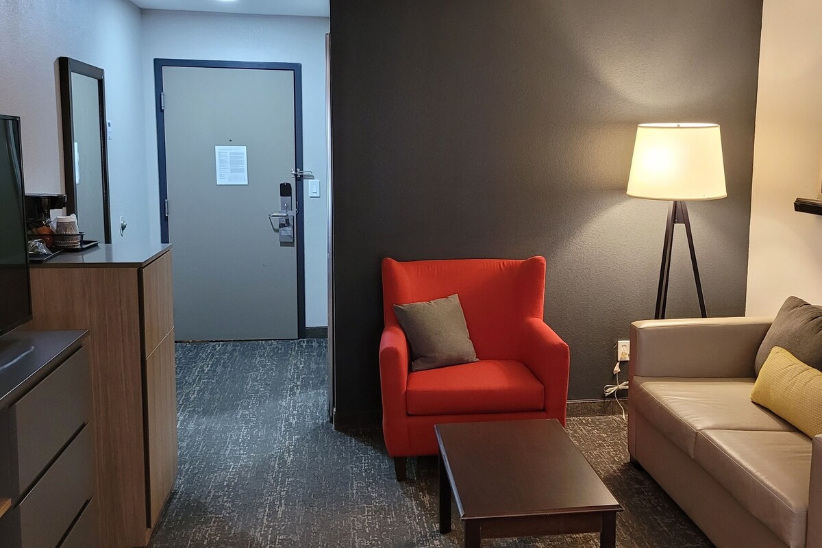 Une chambre d’hôtel avec un fauteuil rouge, un canapé beige, une petite table, une lampe sur pied et un téléviseur posé sur un meuble.