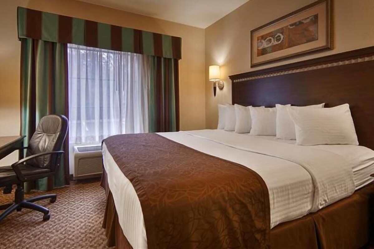 A hotel room with a large bed, a desk, a chair, a window with curtains, and a wall-mounted lamp.