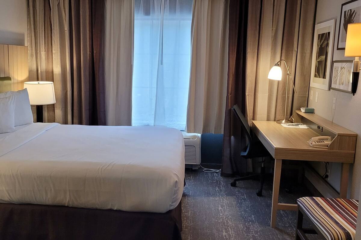 A hotel room with a large bed, two bedside tables with lamps, a desk with a chair, and a window with curtains.