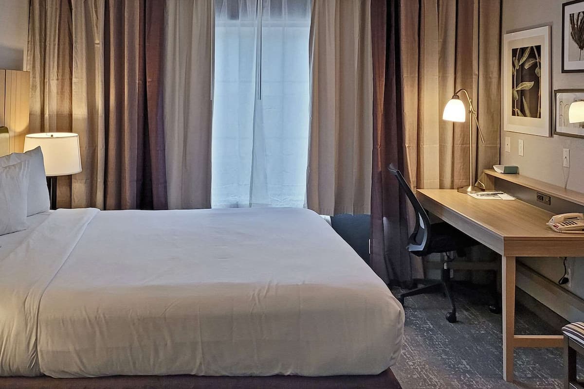 A hotel room with a large bed, a desk with a telephone, a chair, a bench, and a window with curtains.