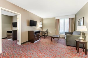 Habitación de hotel con alfombra roja estampada, sofá, escritorio, televisor, lámpara y ventana con cortinas.