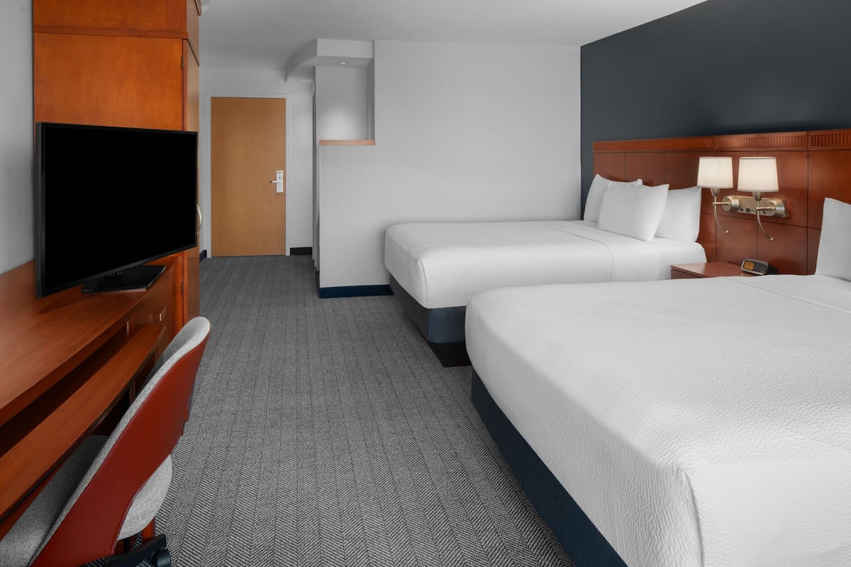 A hotel room with a large bed, a smaller bed, a desk with a TV, and a headboard with lamps.