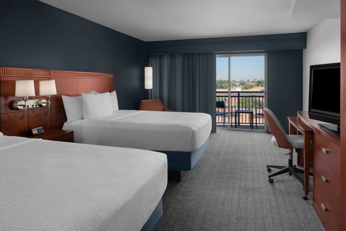 A hotel room with two beds, a desk, a TV, and a balcony with a view.