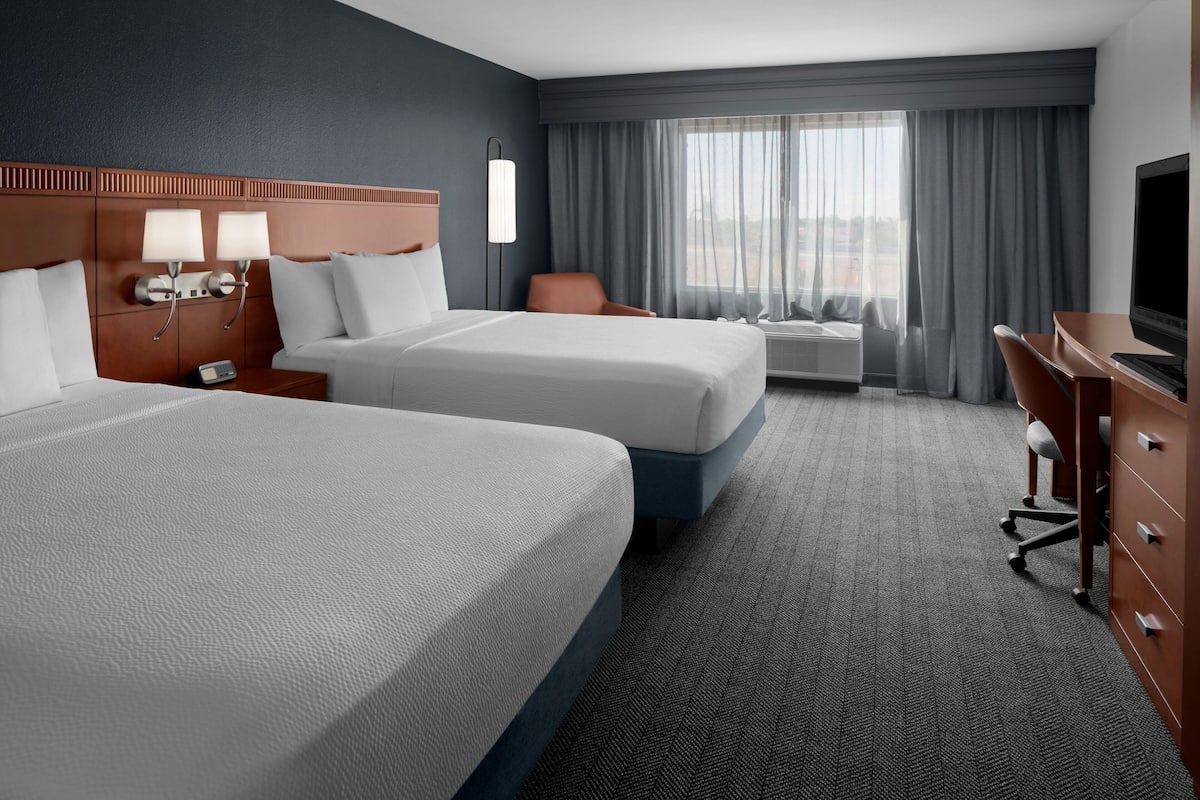 A hotel room with two beds, a desk, a TV, and a window with curtains.