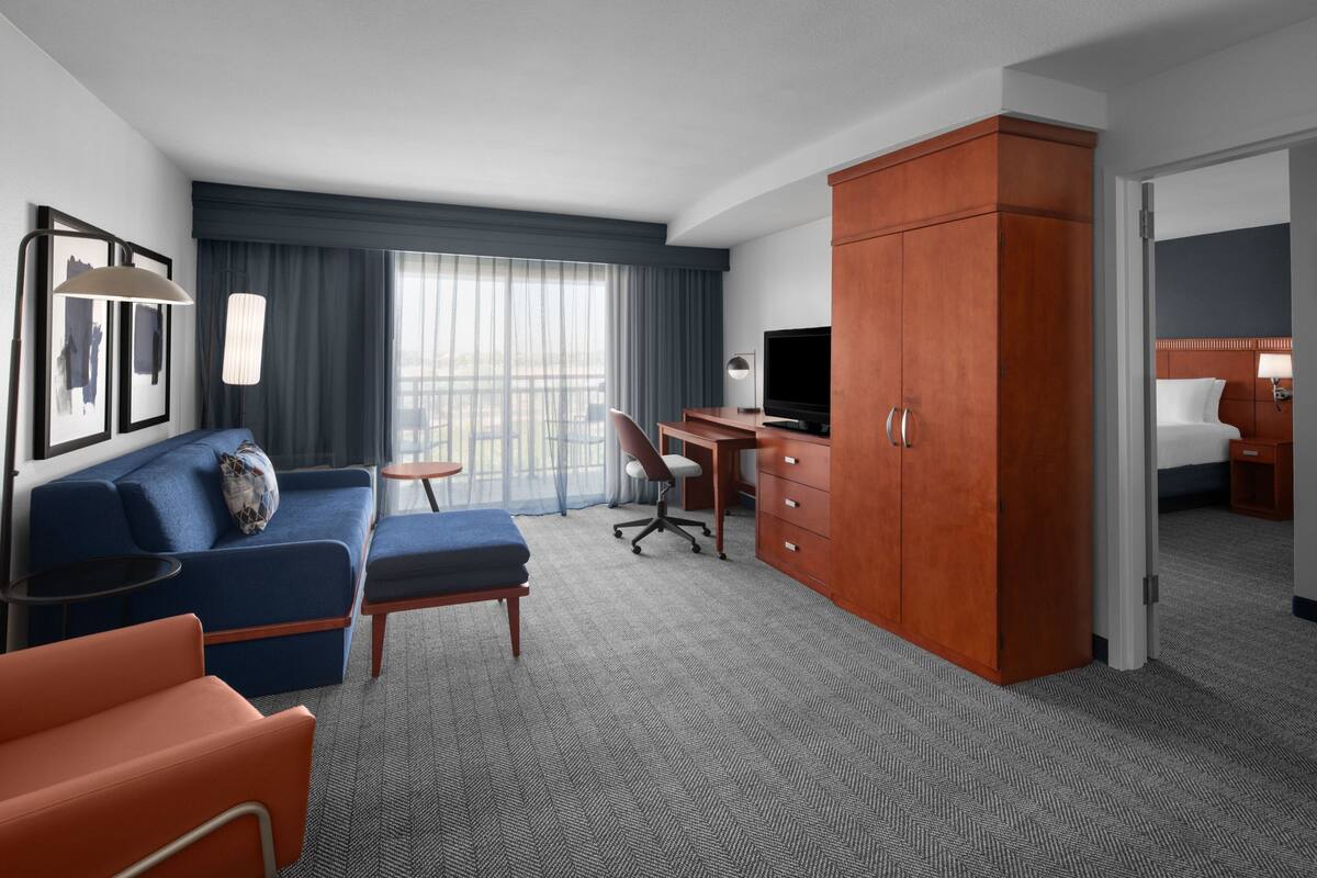 A hotel room with a blue sofa, a wooden wardrobe, a desk with a chair, a television, and a bed with a nightstand.