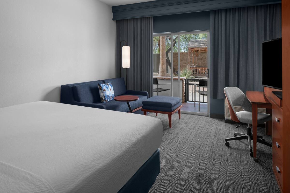 A hotel room with a large bed, a sofa, a desk, and a TV. There is a balcony with a table and chairs.
