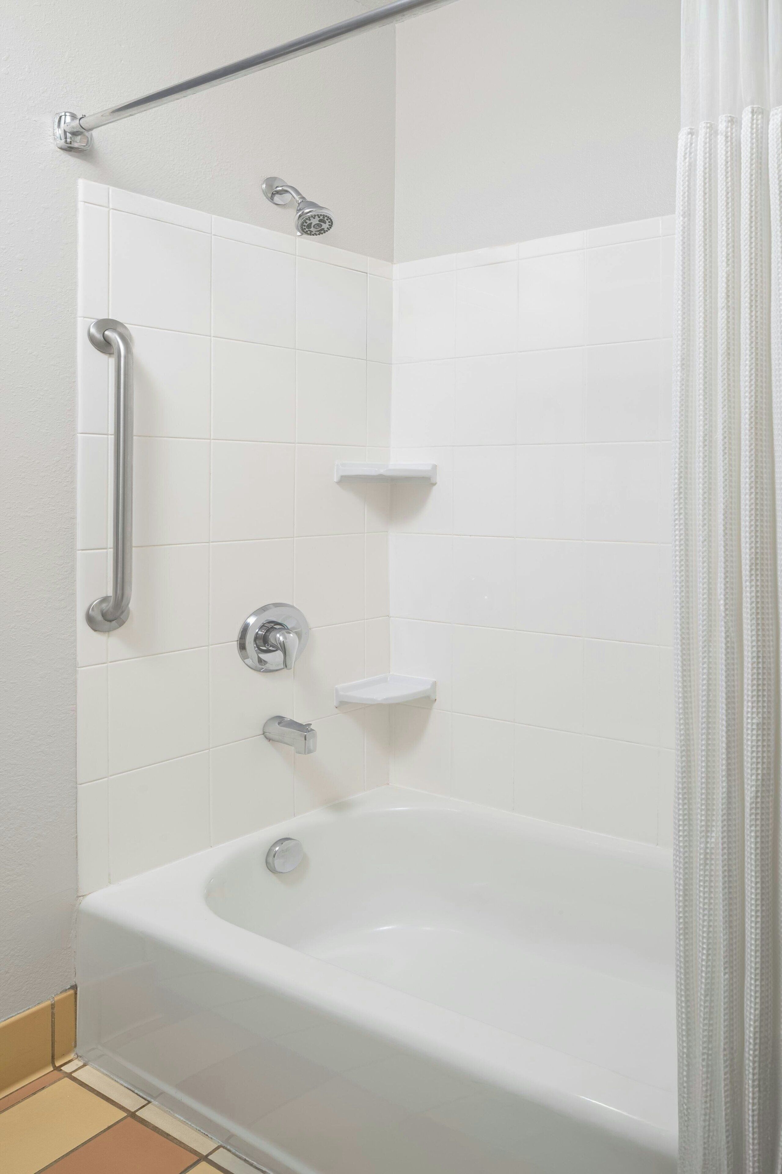 A corner shower with white tiles, a grab bar, a handheld showerhead, and a bathtub.