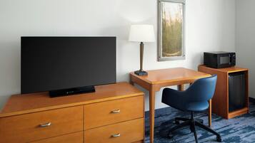 Habitación de hotel con un escritorio de madera, una silla de oficina azul, un televisor de pantalla plana, una lámpara, un microondas y un mueble pequeño.