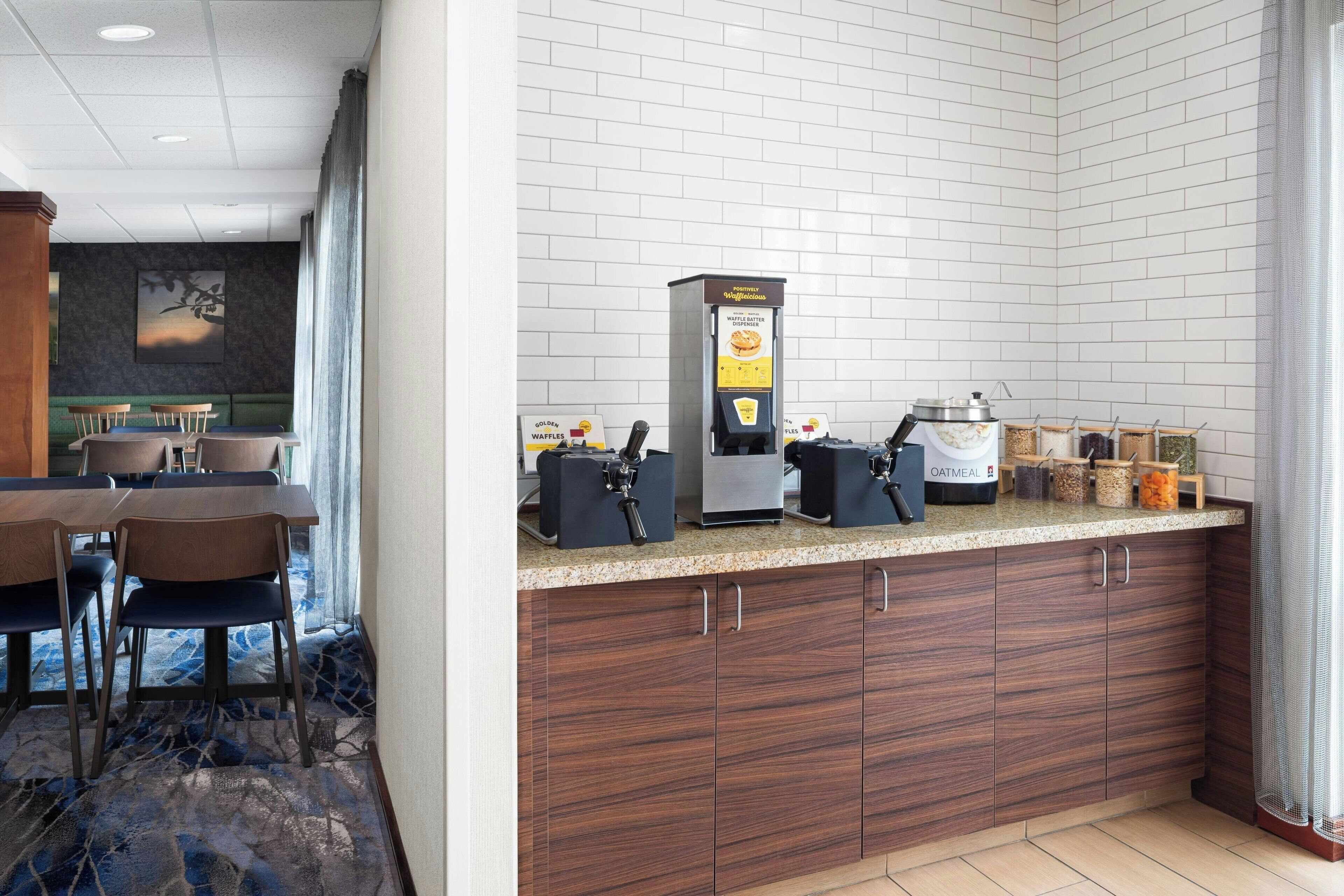 A hotel breakfast area with a waffle batter dispenser, an oatmeal dispenser, and various food containers on the counter.