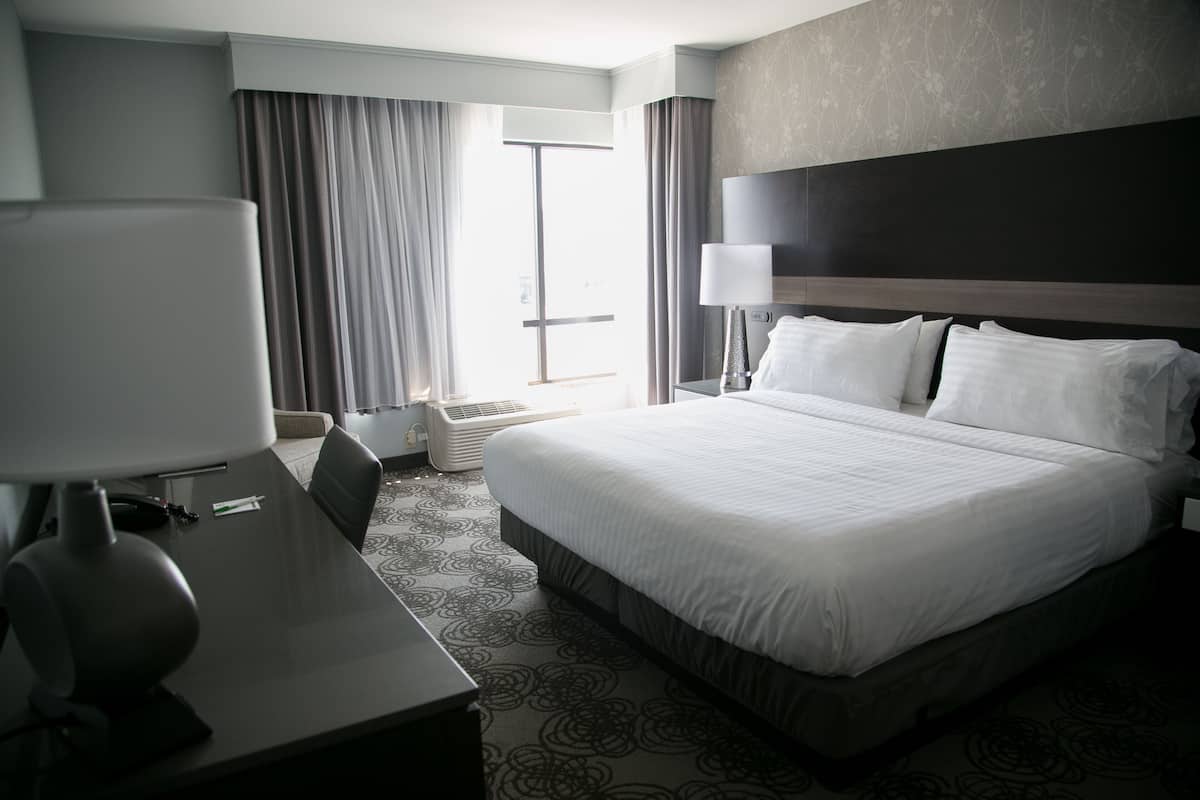 Habitación de hotel con una cama grande, un escritorio con lámpara, una silla y una ventana con cortinas.