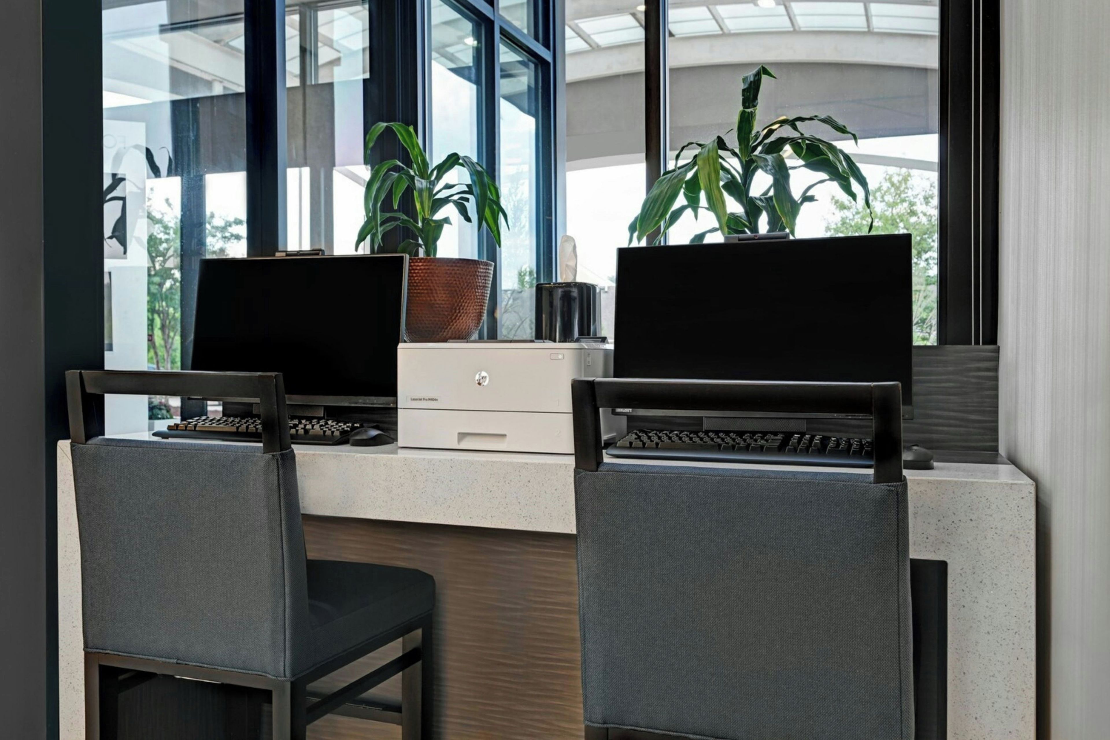 Un espace de travail comprenant deux ordinateurs portables posés sur des bureaux, deux chaises, une petite plante et une fenêtre donnant sur des arbres.