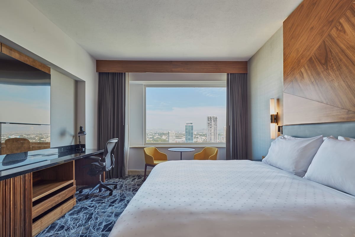 A hotel room with a large bed, a desk with a chair, a small table, and a view of a cityscape through a window.