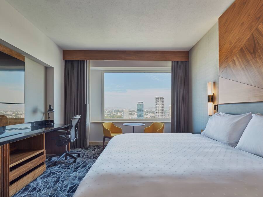 Habitación de hotel con una cama grande, un escritorio con silla, una mesita y vistas a la ciudad por la ventana.