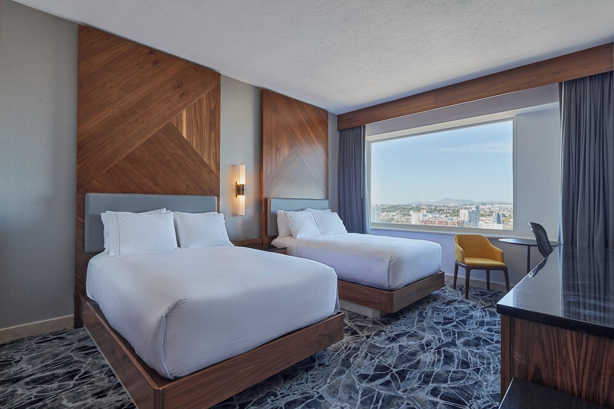 A hotel room with two beds, a desk, and a chair. The room has a large window with a city view.