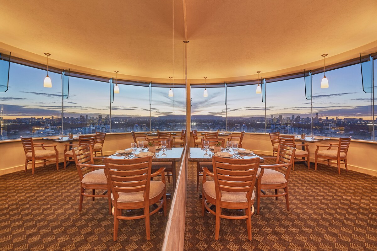 A restaurant with a panoramic city view, round tables, and wooden chairs.
