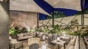 An outdoor seating area with cushioned sofas, chairs, and tables, surrounded by plants and a large umbrella providing shade.