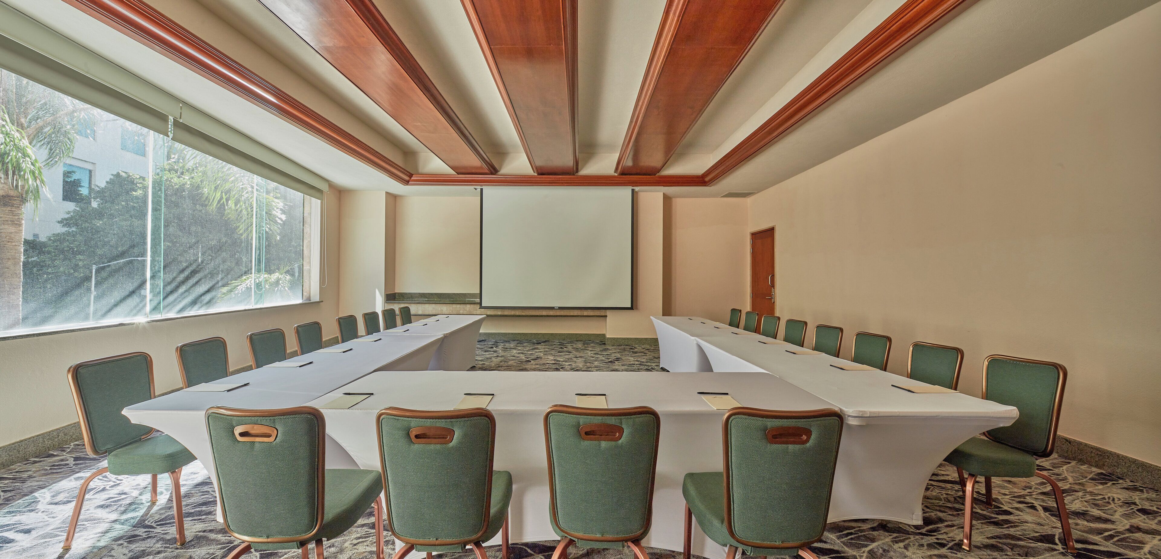 Un salón de conferencias con una larga mesa y sillas tapizadas en verde, ventanas amplias y una pantalla de proyector.