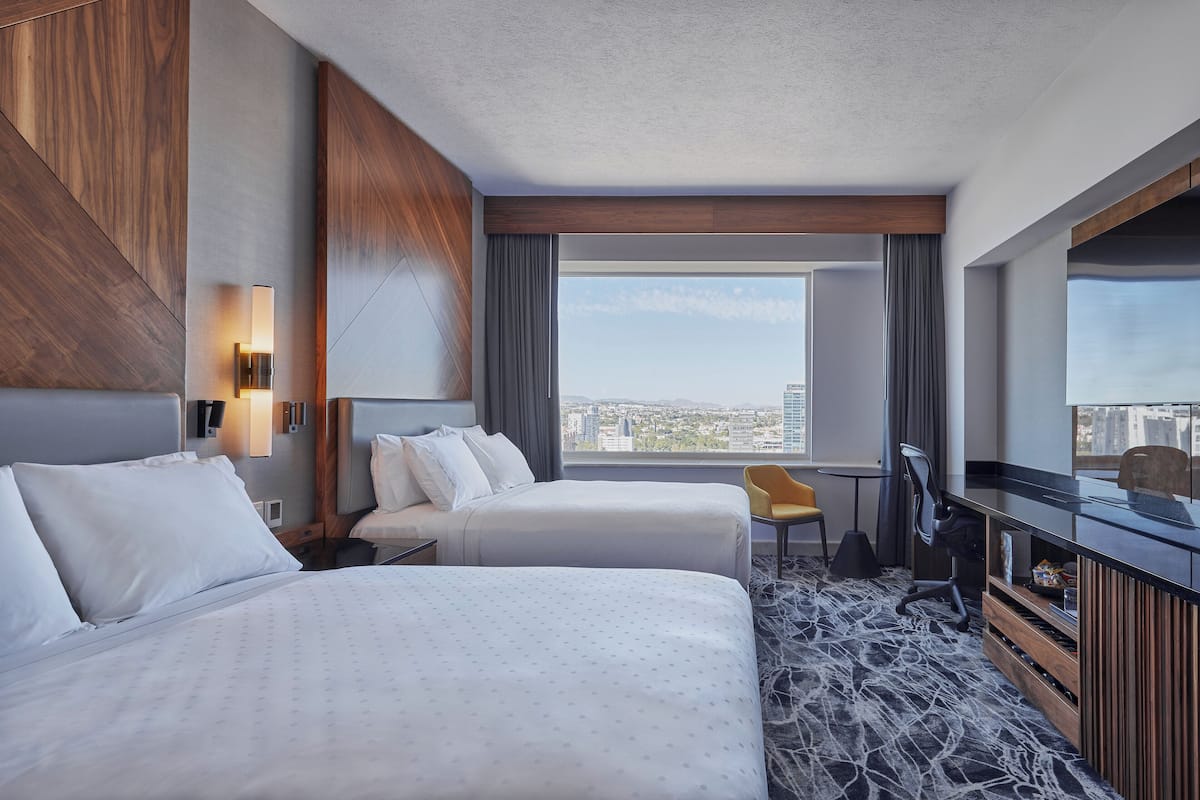 A hotel room with two beds, a desk, a chair, a TV, and a large window offering a city view.