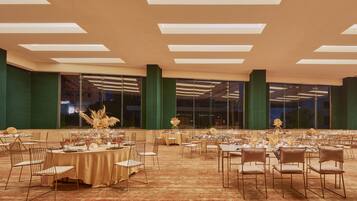 Uma sala de banquetes com mesas redondas preparadas para refeições, amplas janelas e teto alto com iluminação embutida.