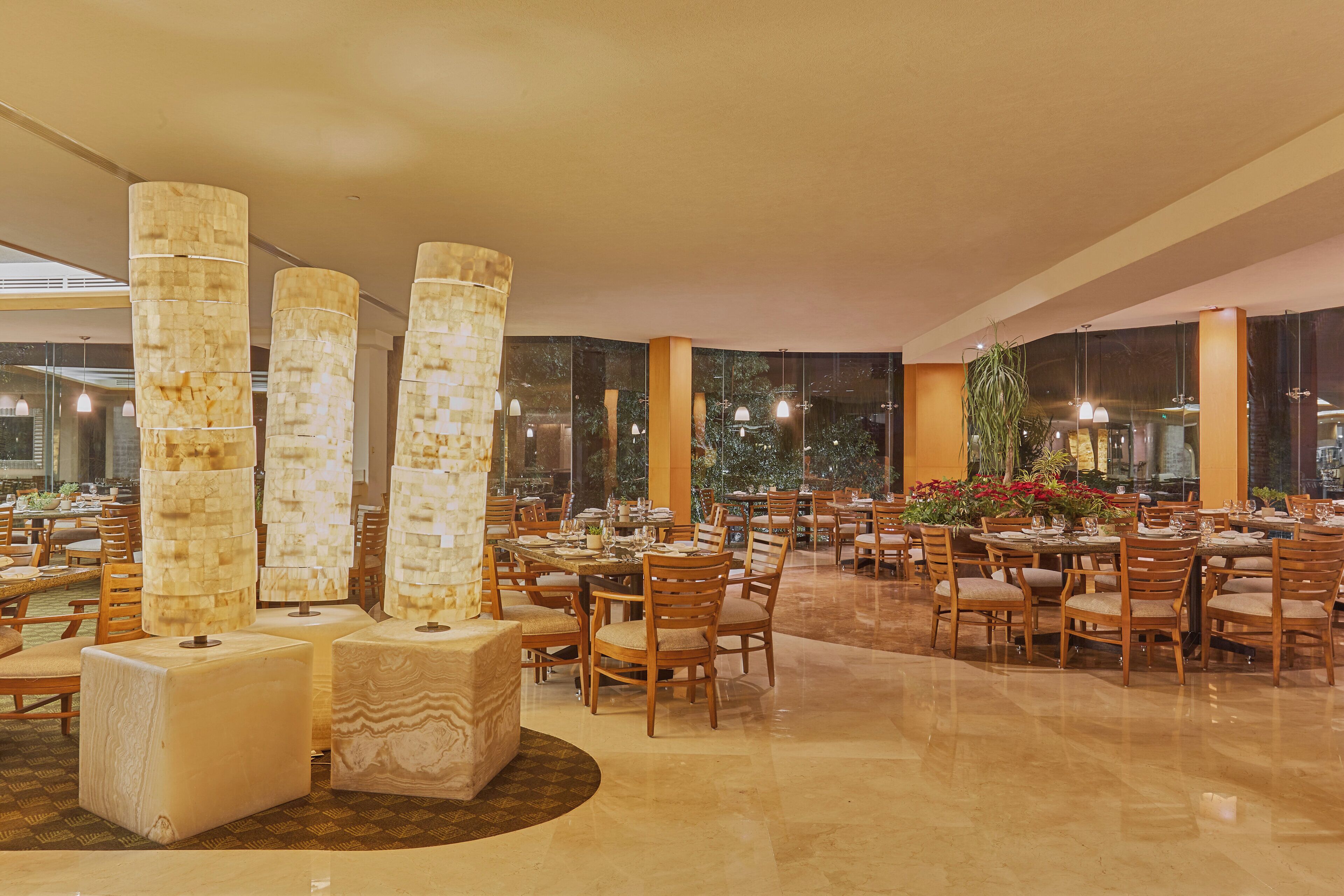 A spacious dining area with multiple tables and chairs, large decorative columns, and a view of the outdoors through large windows.