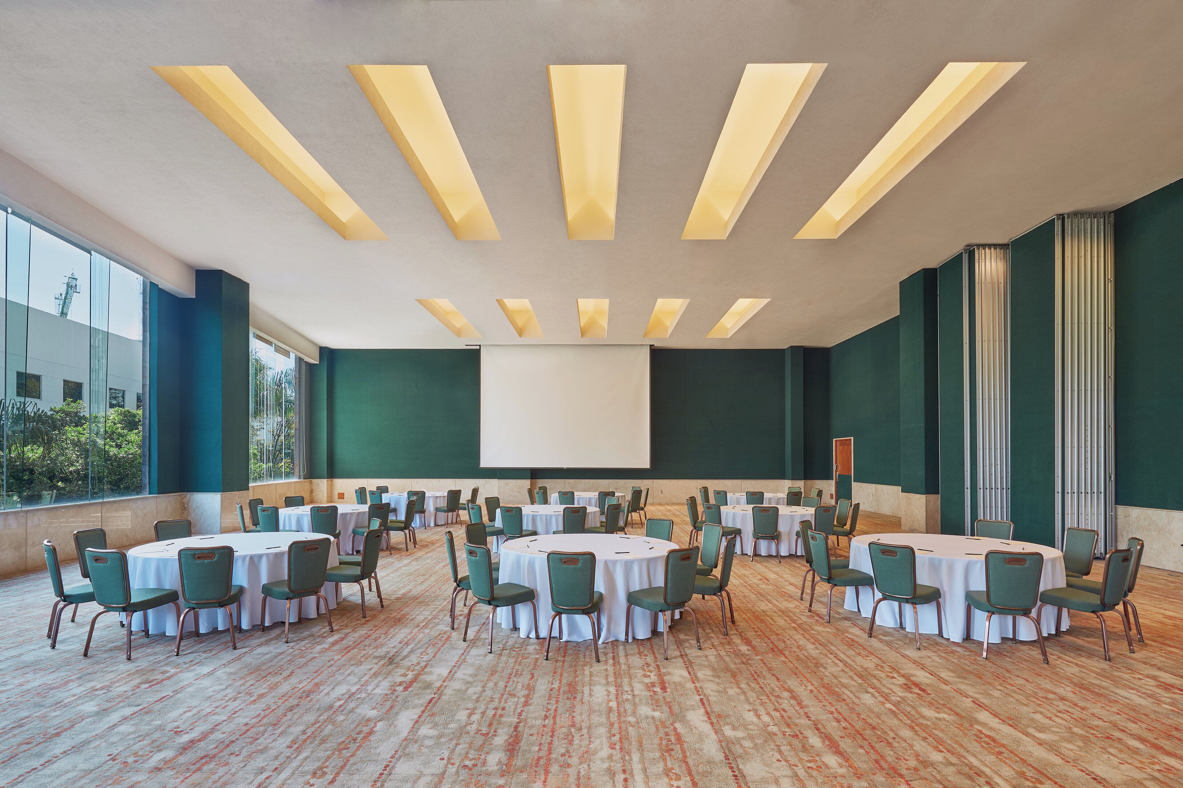 Un salón de conferencias con varias mesas redondas y sillas, ventanas amplias y una pantalla de proyector.