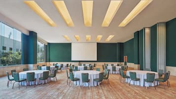 Un salón de conferencias con varias mesas redondas y sillas, ventanas amplias y una pantalla de proyector.