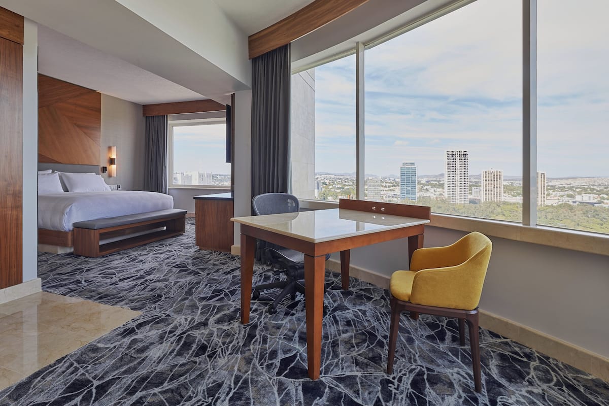 A hotel room with a bed, a desk, a chair, and a large window offering a city view.