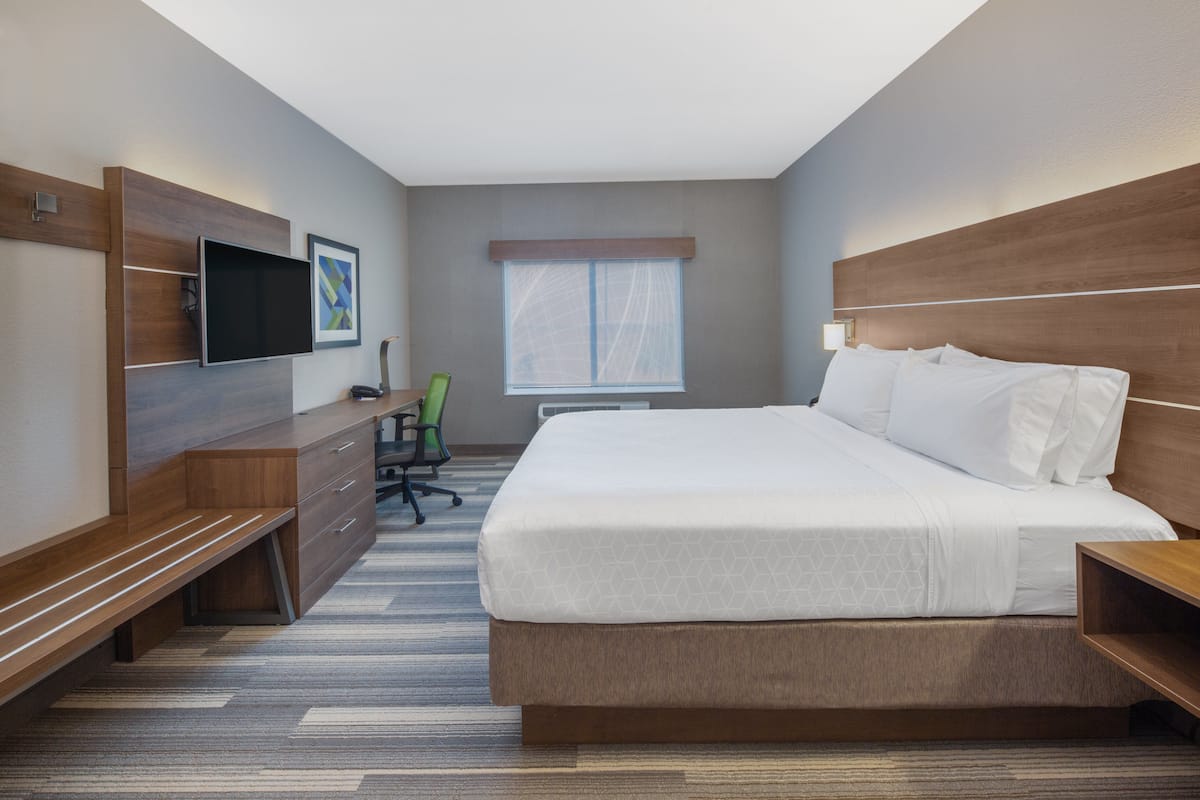 A modern hotel room with a large bed, a flat-screen TV mounted on the wall, a desk with a chair, and a window with blinds.