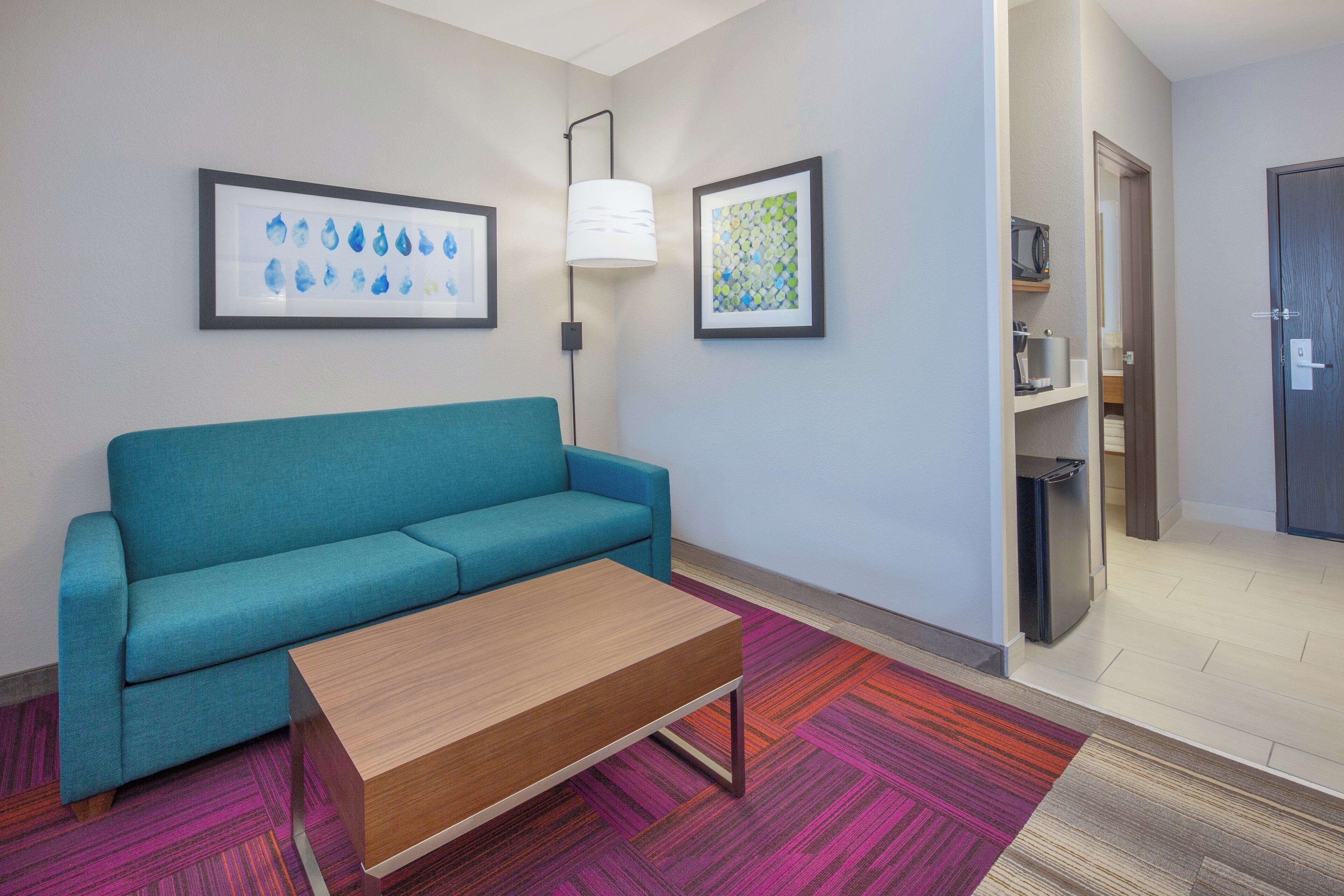 A hotel room with a teal sofa, a wooden coffee table, a colorful rug, a microwave, and a refrigerator.