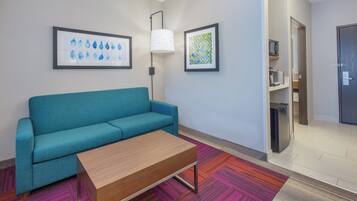 A hotel room with a teal sofa, a wooden coffee table, a colorful rug, a microwave, and a refrigerator.