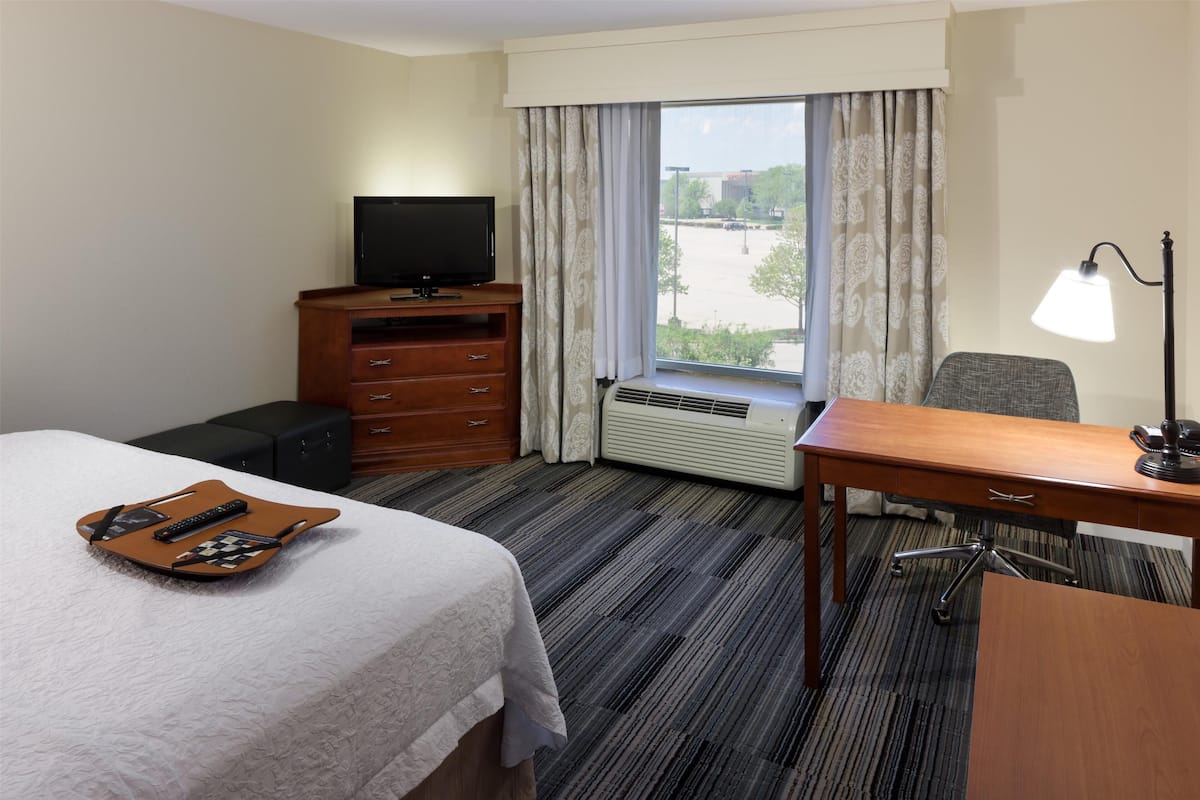 A hotel room with a bed, a desk with a lamp, a TV, a dresser, and a window with curtains.