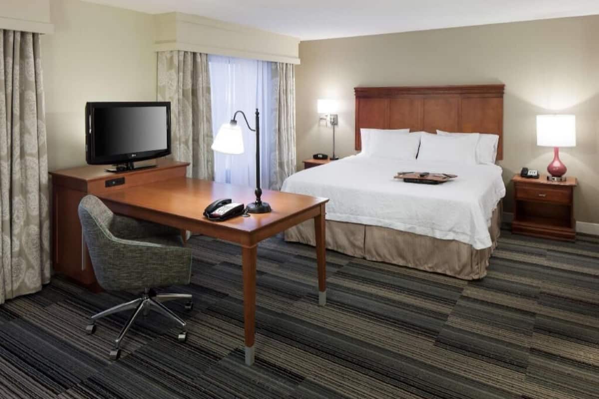 A hotel room with a bed, a desk with a television, a chair, a lamp, and a bedside table.