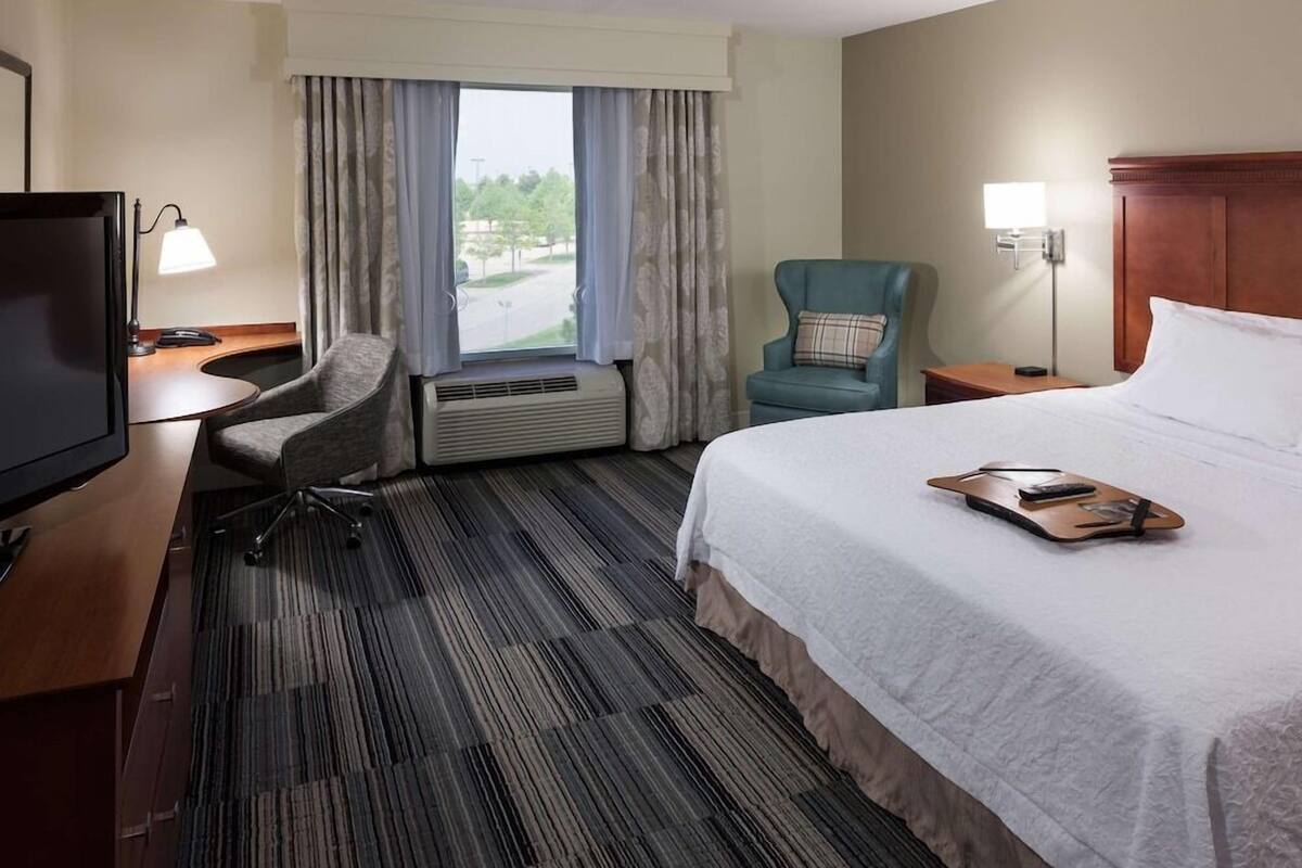 A hotel room with a bed, a desk with a TV, a chair, and a window with curtains.