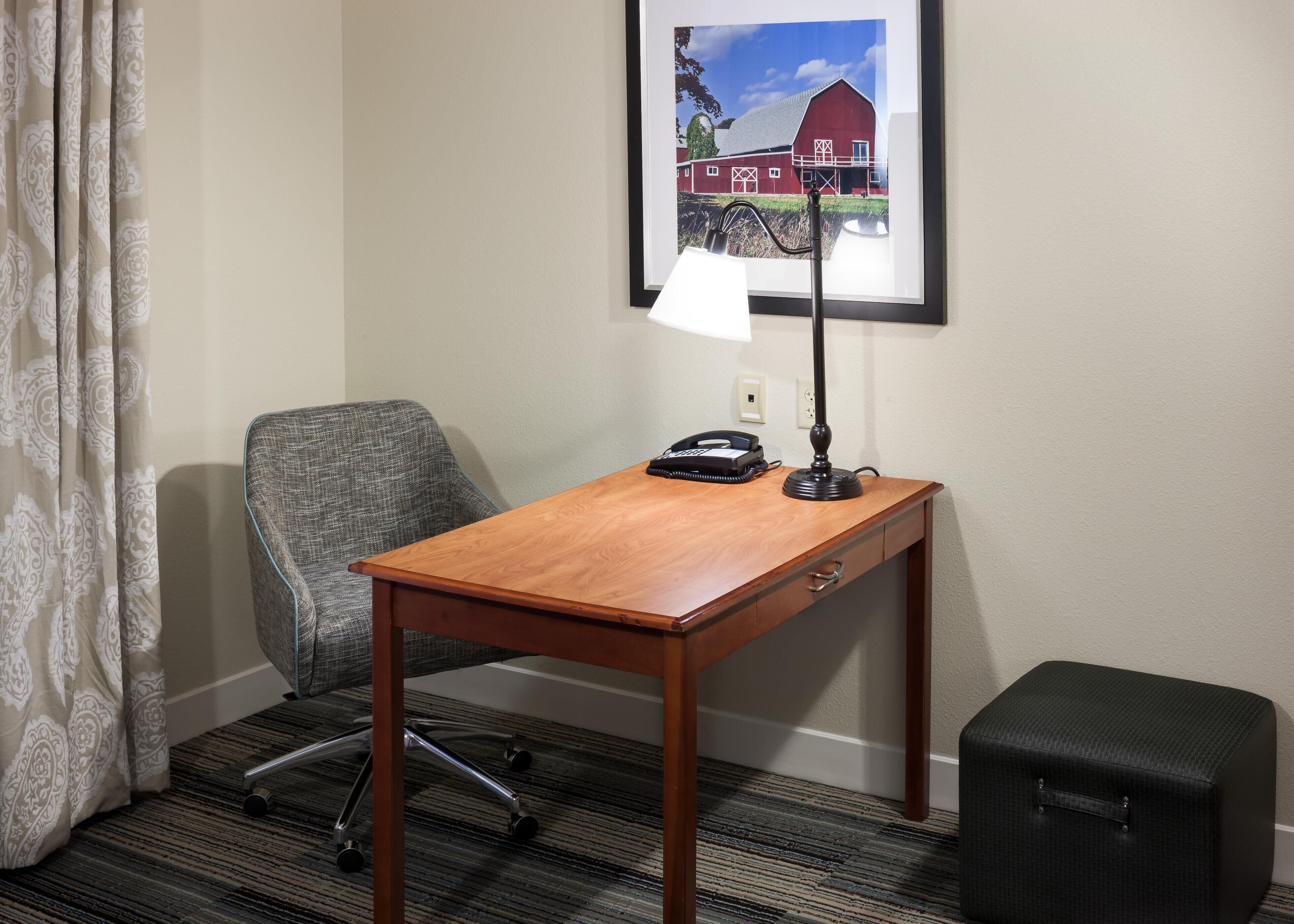 A hotel room with a wooden desk, a gray office chair, a black telephone, a desk lamp, a framed picture of a barn, and a black suitcase.