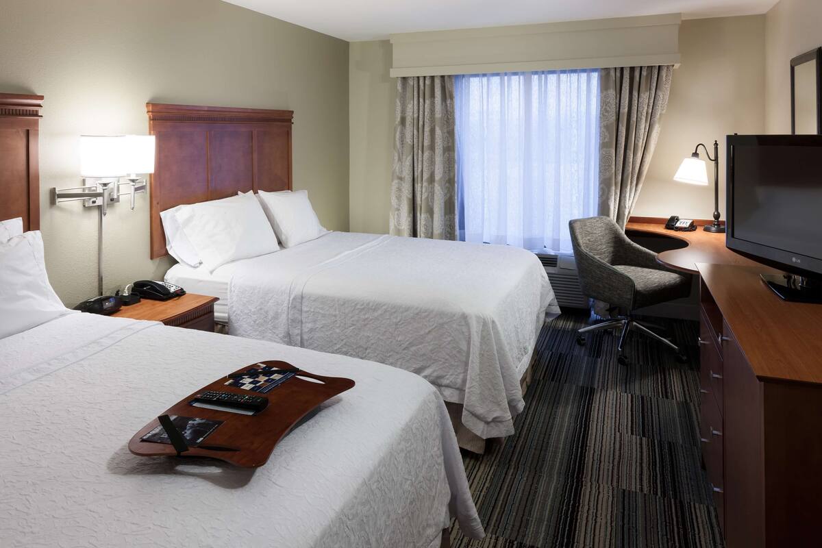 A hotel room with two beds, a desk with a chair, a television, and a window with curtains.