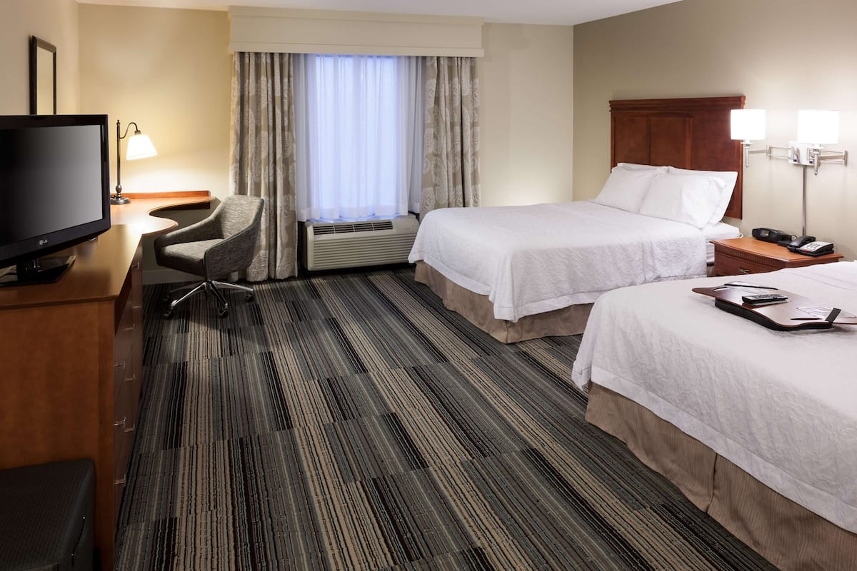 A hotel room with a flat-screen TV, two beds, a desk, a chair, and a window with curtains.