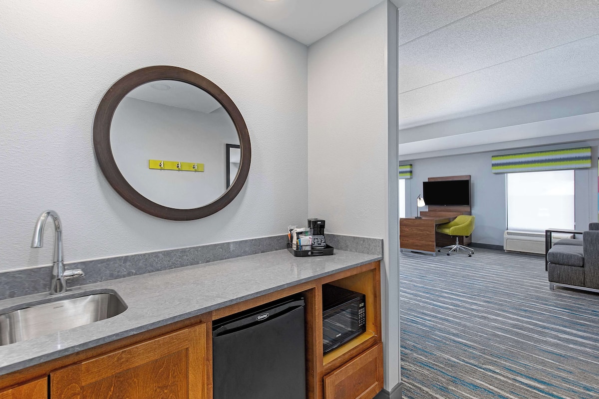 A hotel room with a sink, mirror, coffee maker, microwave, and a seating area with a TV and windows.