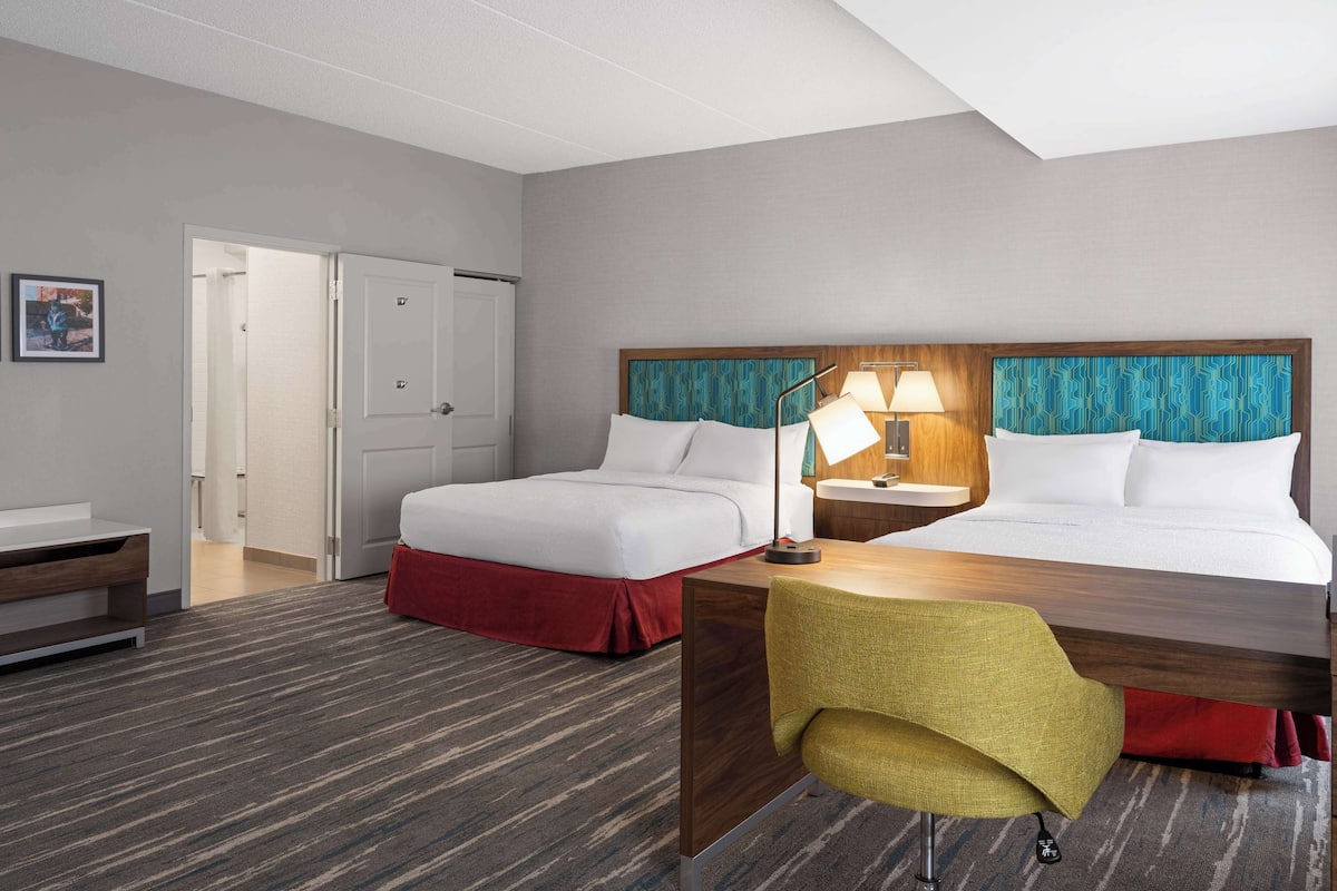 A hotel room with two beds, a desk, a chair, and a lamp.