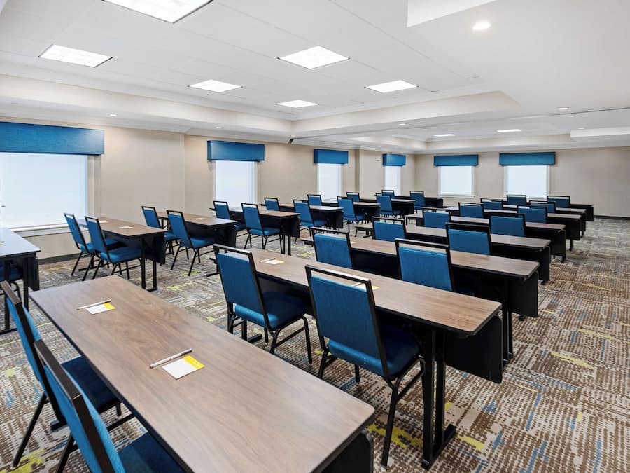 A conference room with rows of tables and chairs, large windows with blinds, and a carpeted floor.