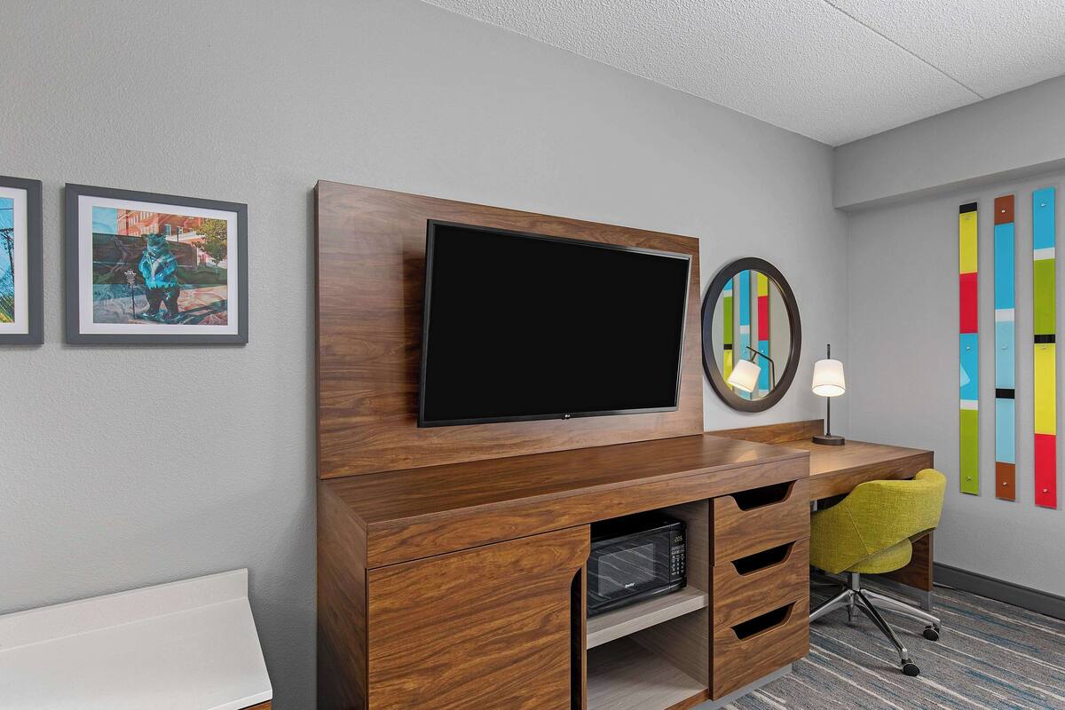 A hotel room with a wooden desk, a flat-screen TV, a microwave, a yellow chair, and artwork on the walls.