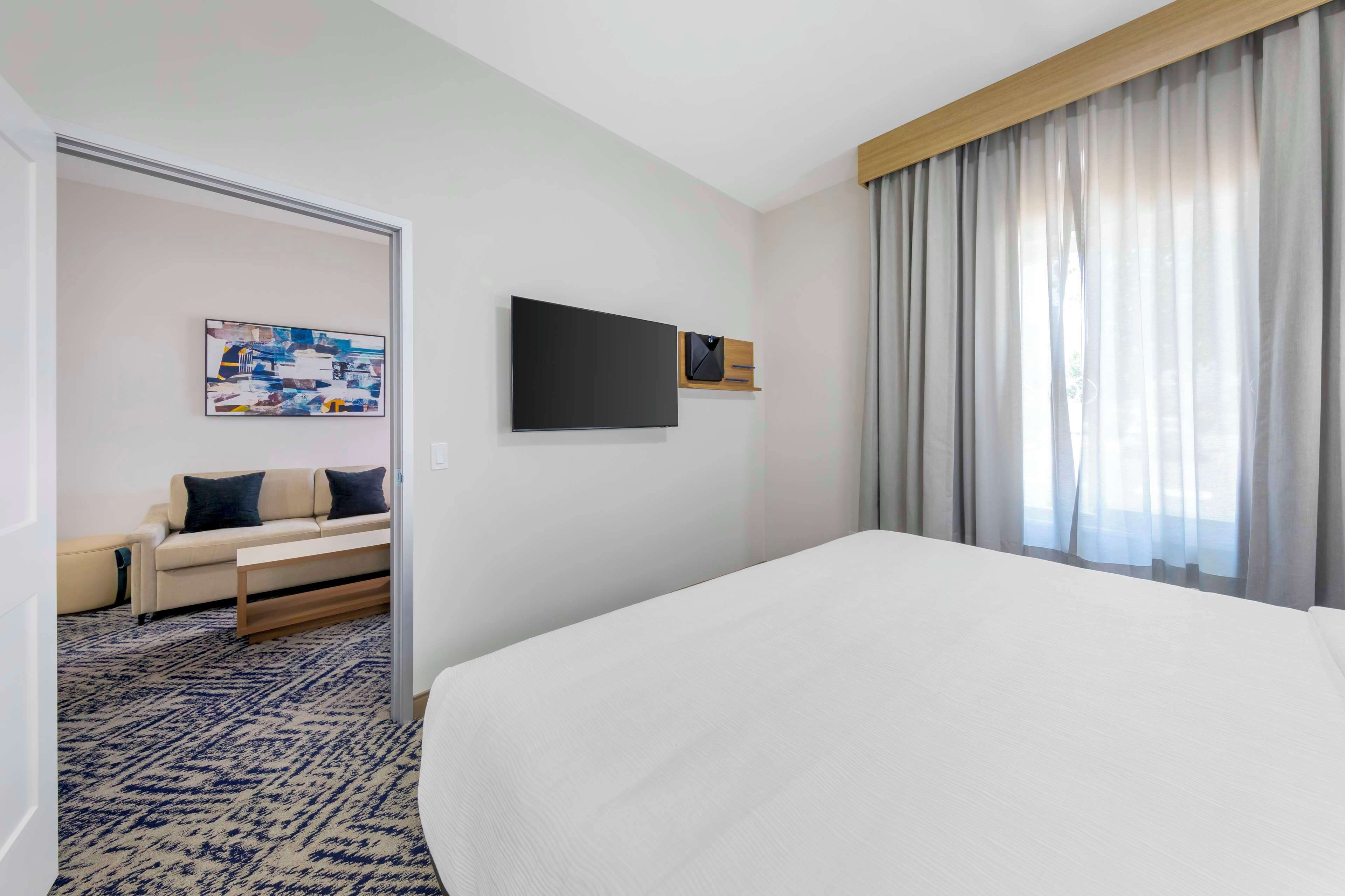 A hotel room with a large bed, a flat-screen TV mounted on the wall, a sofa in the corner, and a large window with curtains.