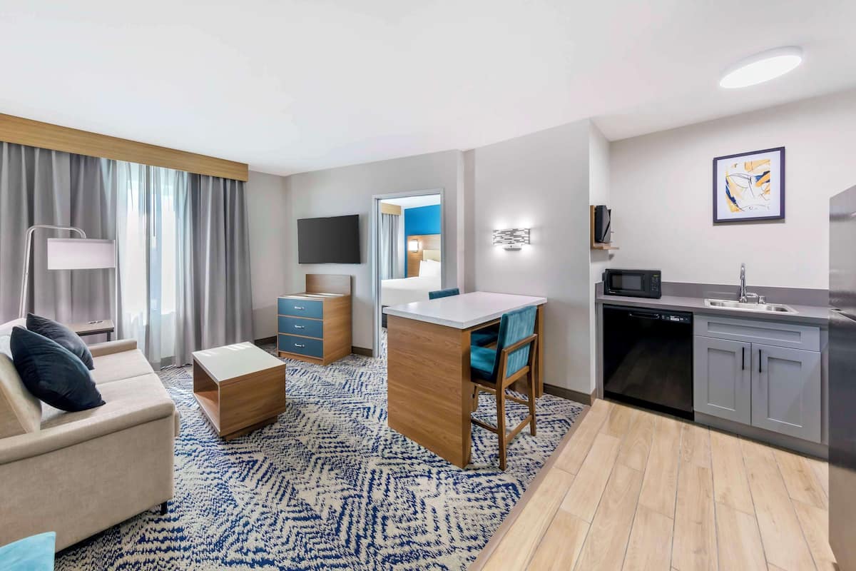 A modern hotel room with a living area, a small kitchenette, a desk, and a large window with curtains.