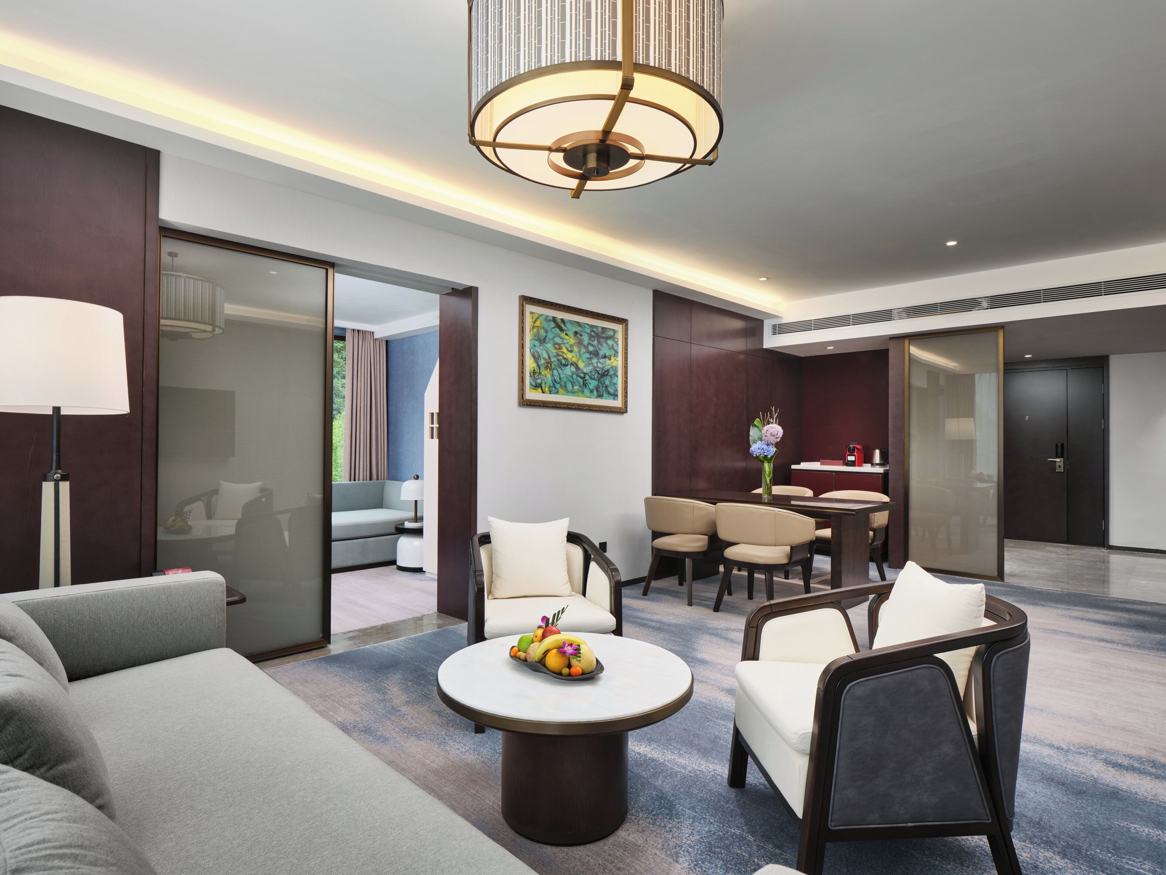 Habitación de hotel con sofá, mesa de comedor con sillas, frutero, lámpara y un cuadro en la pared.