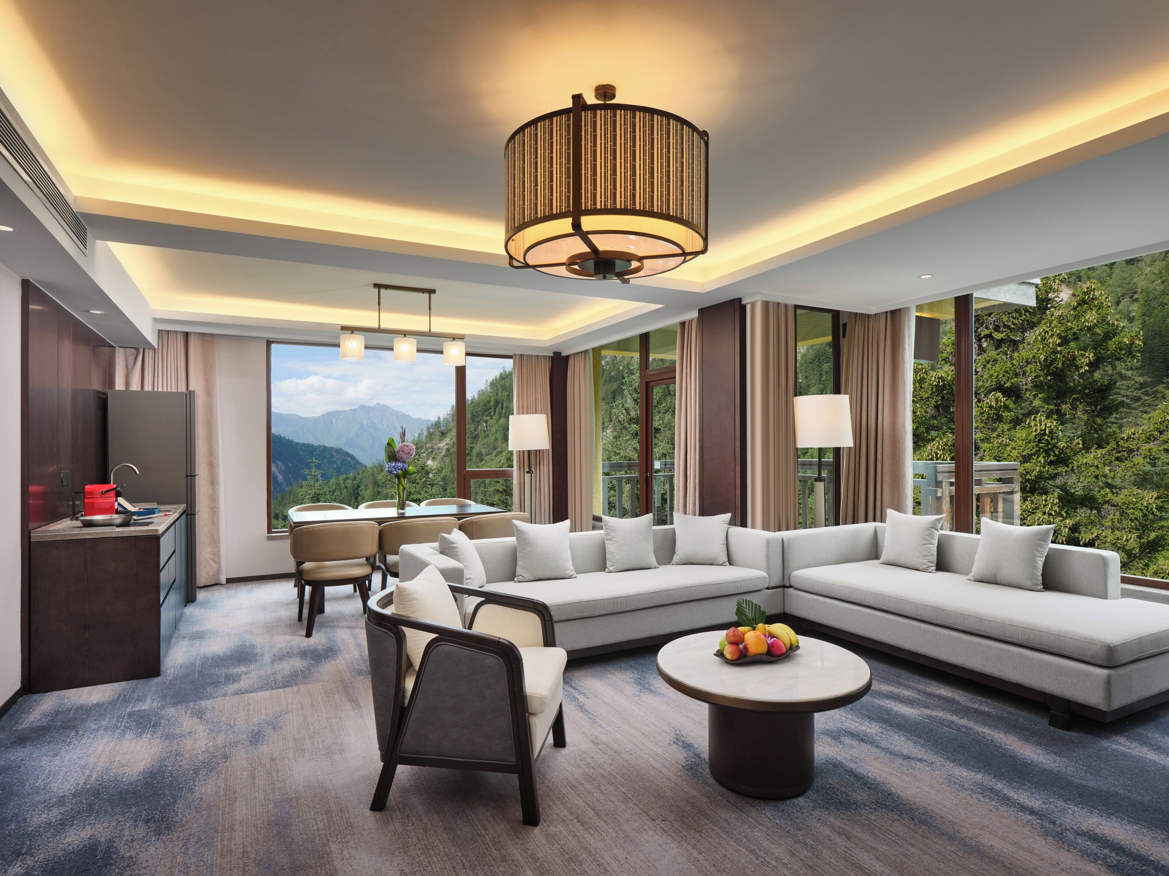 A modern hotel room with a large sofa, dining table, and chairs, a kitchen area, and a view of the mountains.