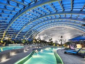 Uma grande piscina interior com teto de vidro, rodeada por espreguiçadeiras e palmeiras.
