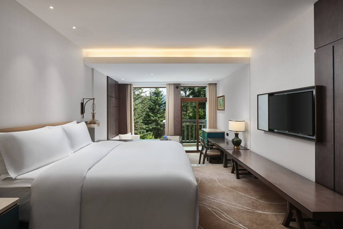 Une chambre d’hôtel moderne dotée d’un grand lit, d’un bureau, d’une télévision à écran plat et d’un balcon offrant une vue sur les arbres.