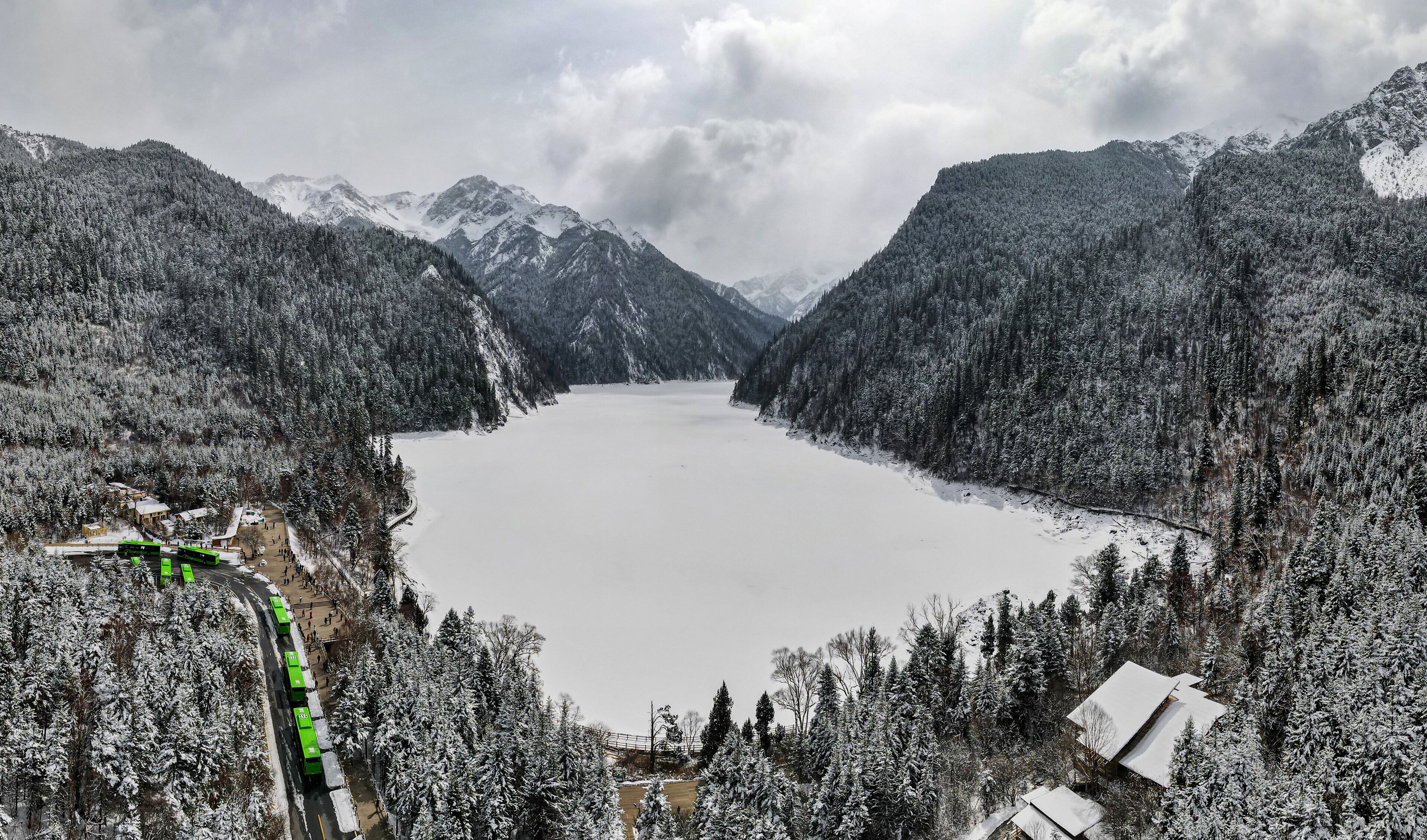 A snow-covered lake surrounded by forested mountains, with a green train on tracks to the left.