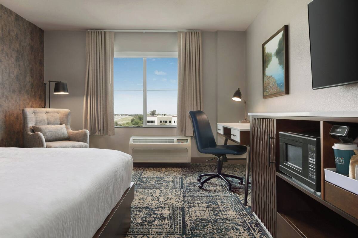 A hotel room with a large bed, a desk, a chair, a microwave, and a coffee cup.