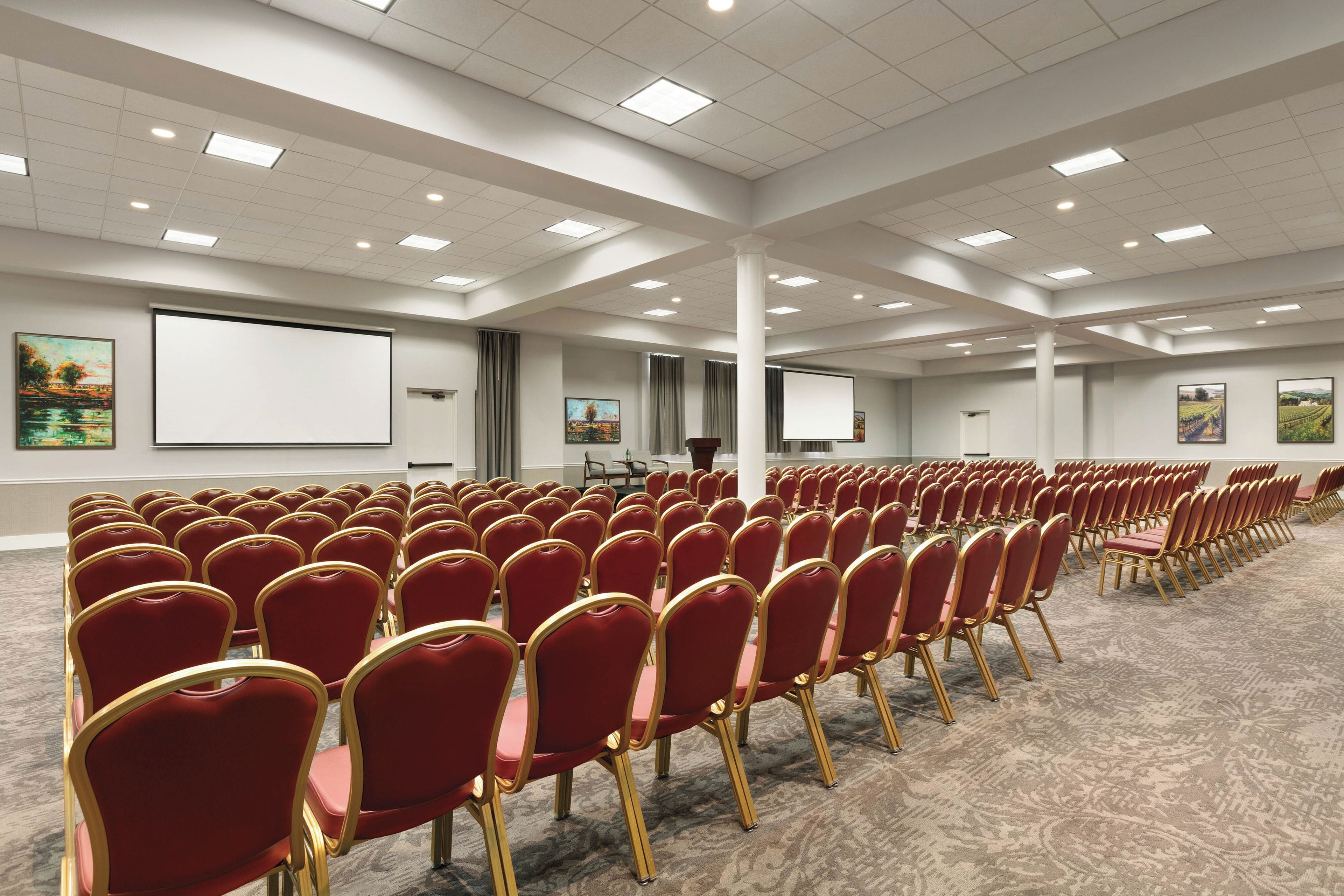 Konferenssihuone, jossa on riveissä punaisia tuoleja, suuri näyttö ja matolla peitetty lattia.