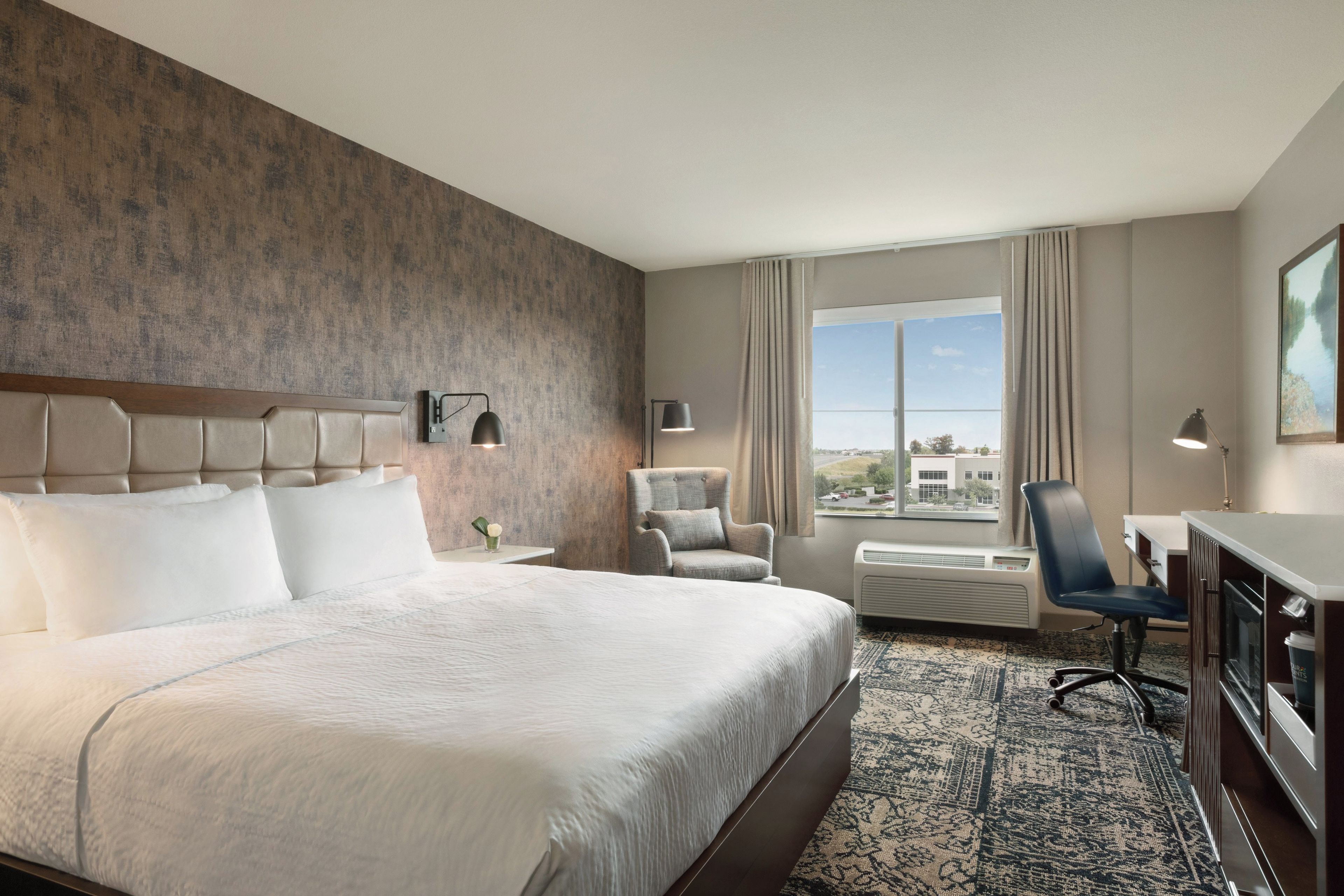 Une chambre d’hôtel avec un grand lit, un bureau, une chaise, une télévision et une fenêtre donnant sur un beau paysage.